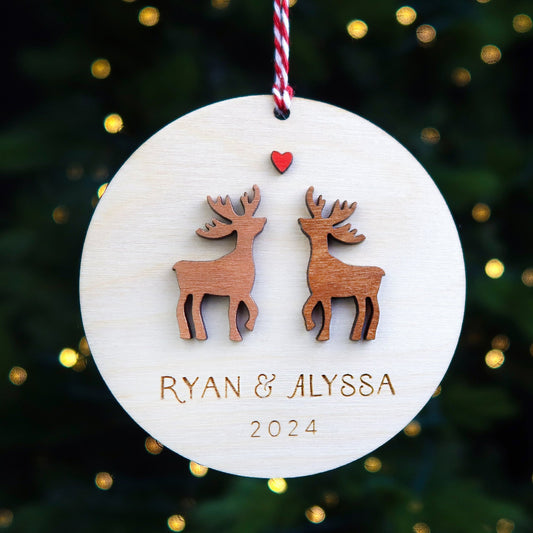 A personalized couple's Christmas ornament with a birch wood disc, hand-painted reindeer shapes, and names and a date engraved on it. The ornament is hanging from twine, with a red and white Christmas tree in the background.