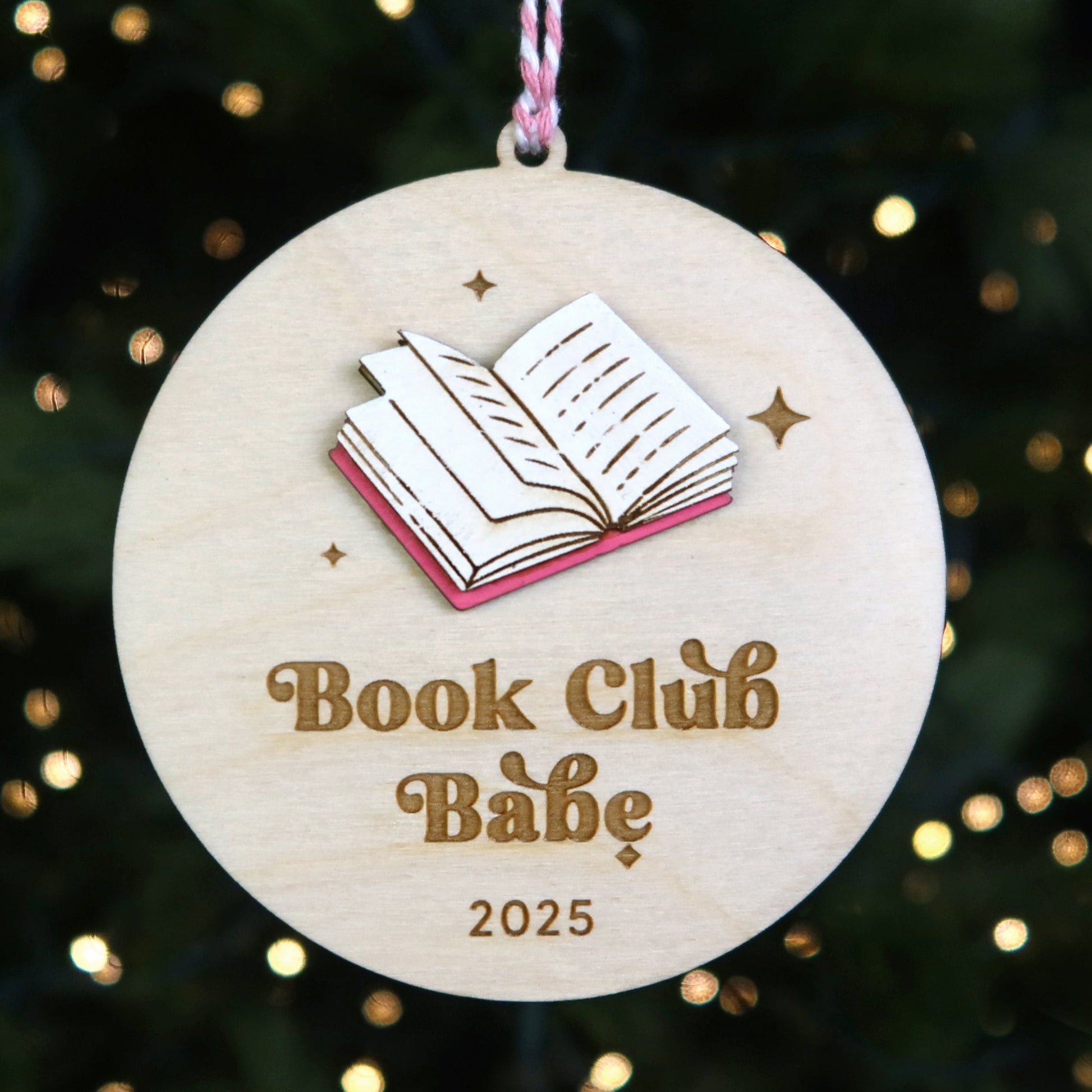 Wooden ornament with book design and 'Book Club Babe 2025' text on a Christmas tree background.