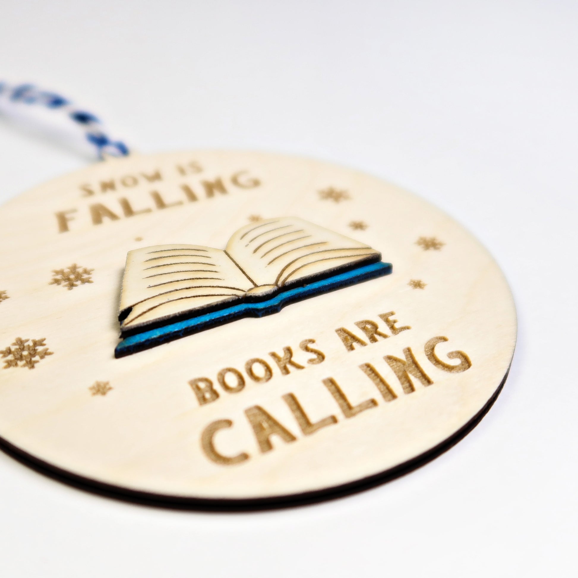 Wooden ornament with book illustration and text 'Now is falling, books are calling' on a white background