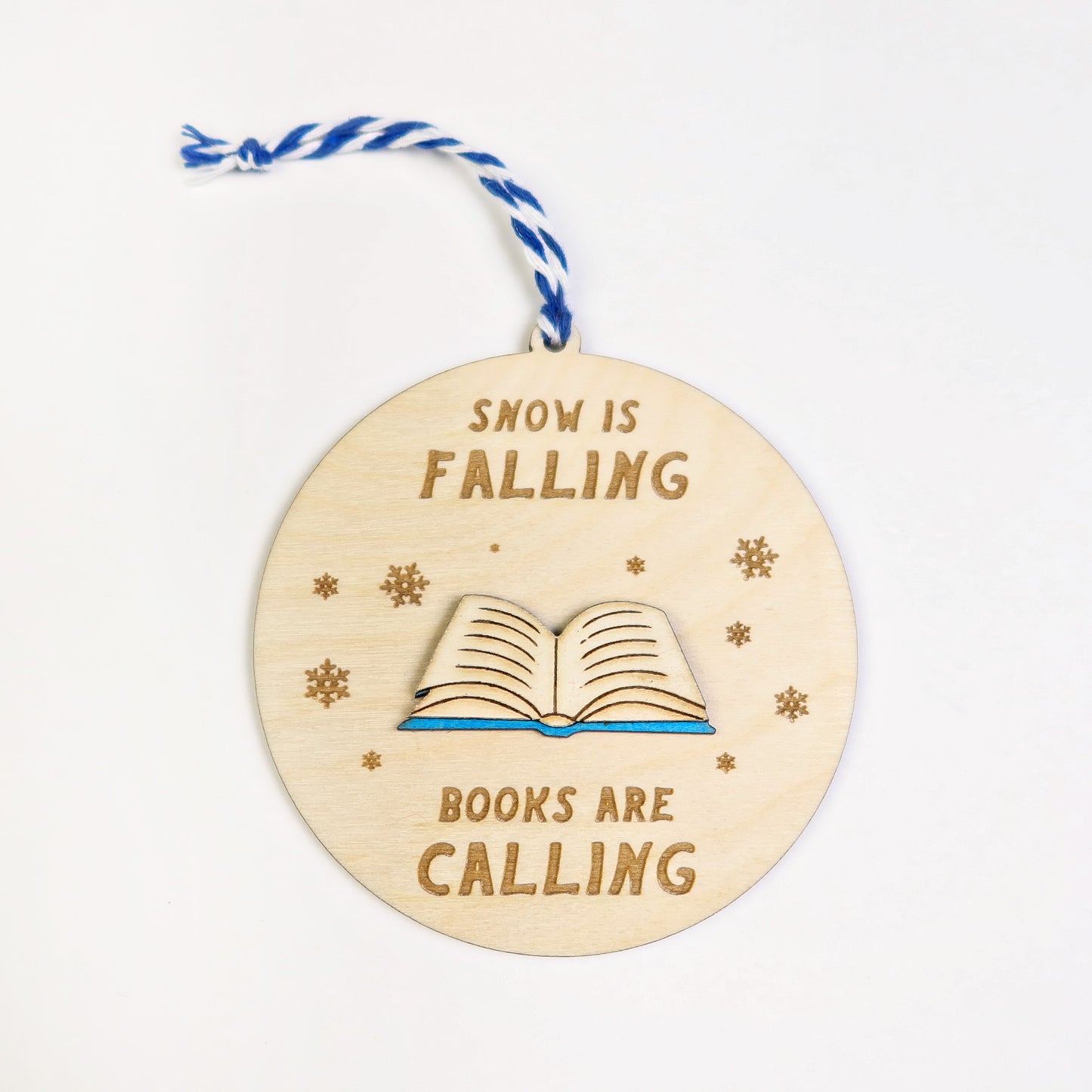 Wooden ornament with 'Snow is falling, books are calling' text and a book illustration on a white background.