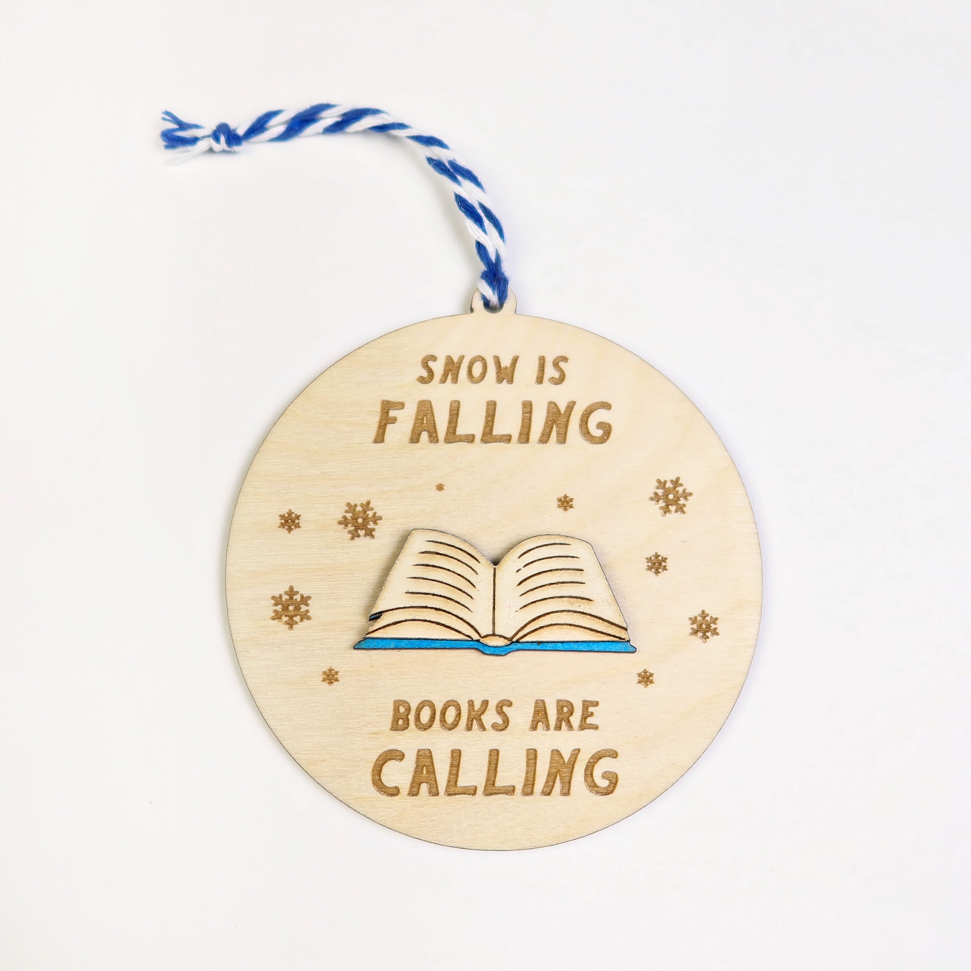 Wooden ornament with 'Snow is falling, books are calling' text and a book illustration on a white background.