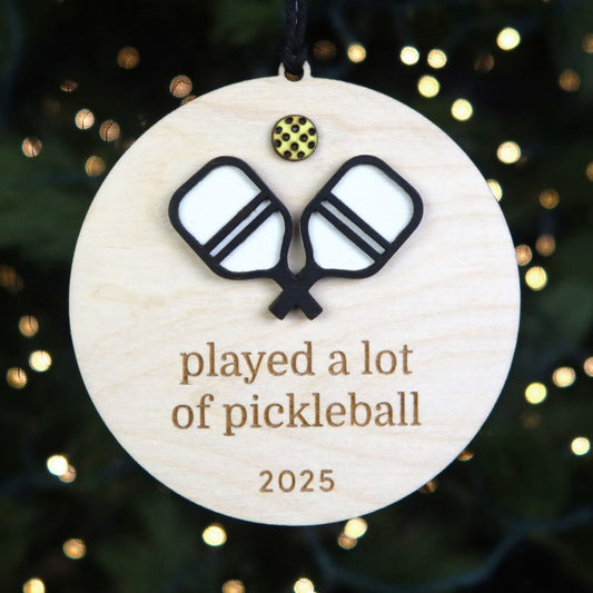 Round wooden ornament with pickleball design and text 'played a lot of pickleball 2025' against a festive background.