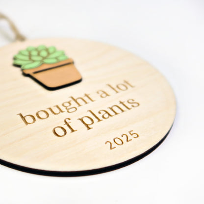 Wooden tag with a potted plant illustration and text 'bought a lot of plants 2025' on a white background