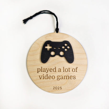 Wooden ornament with a black game controller silhouette and text 'played a lot of video games 2025' on a white background.