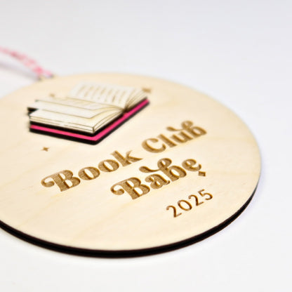 Round wooden tag with 'Book Club Babe 2025' text and book illustration on a white background