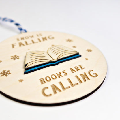 Wooden ornament with book illustration and text 'Now is falling, books are calling' on a white background