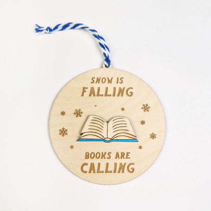 Wooden ornament with 'Snow is falling, books are calling' text and a book illustration on a white background.