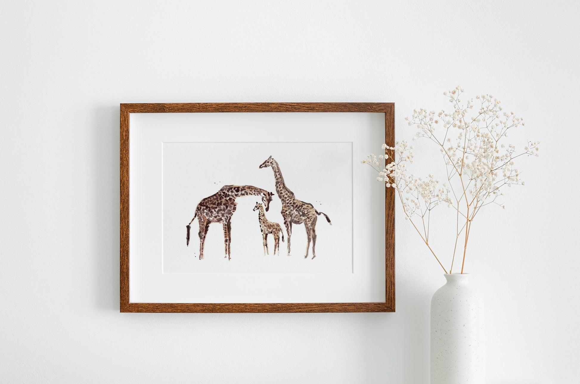 Framed artwork of giraffes on a white wall with a vase of dried flowers.